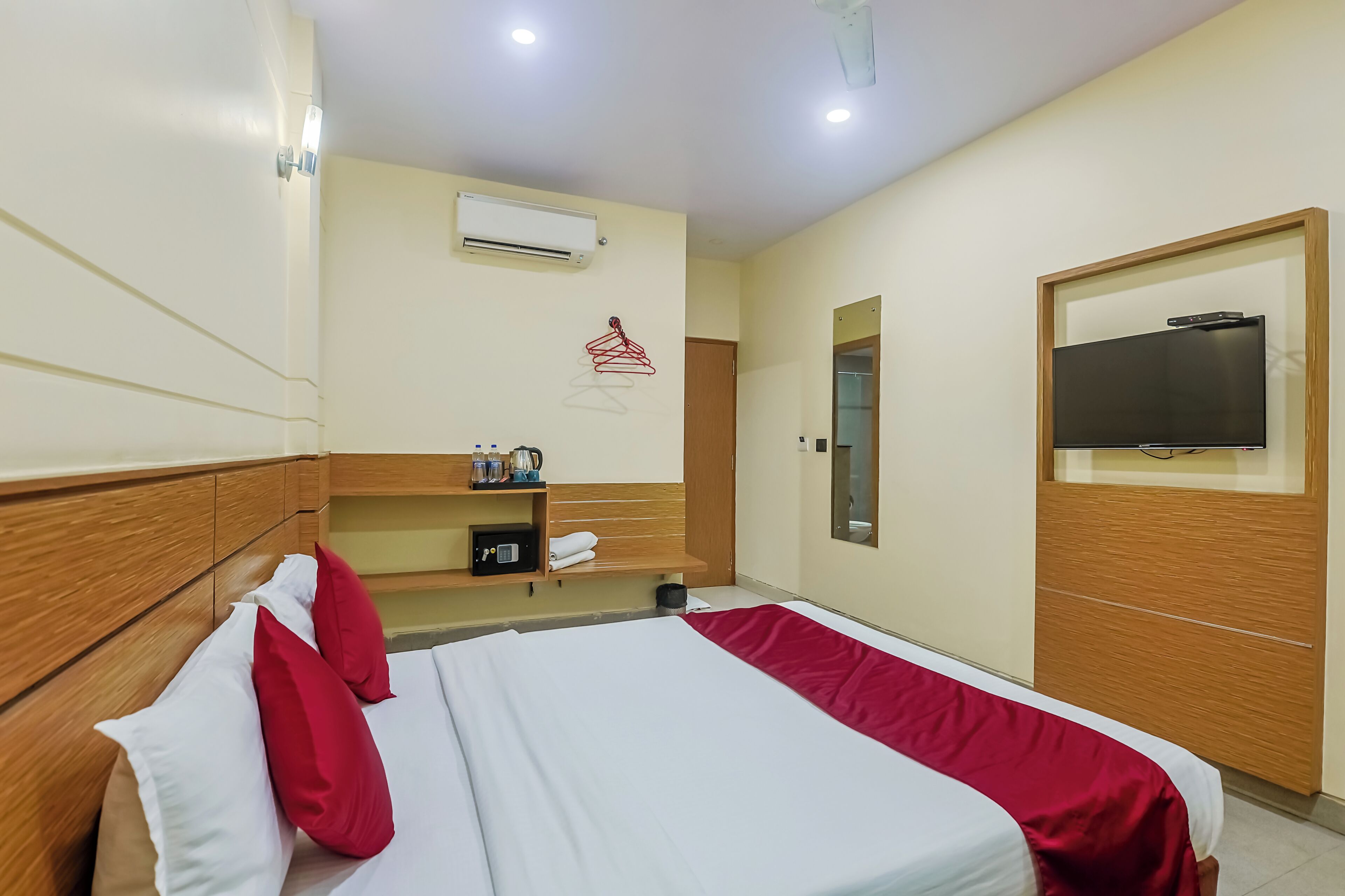 A hotel room with a wooden headboard, a bed with white and red bedding, a TV mounted on the wall, a small shelf with toiletries, and an air conditioning unit.