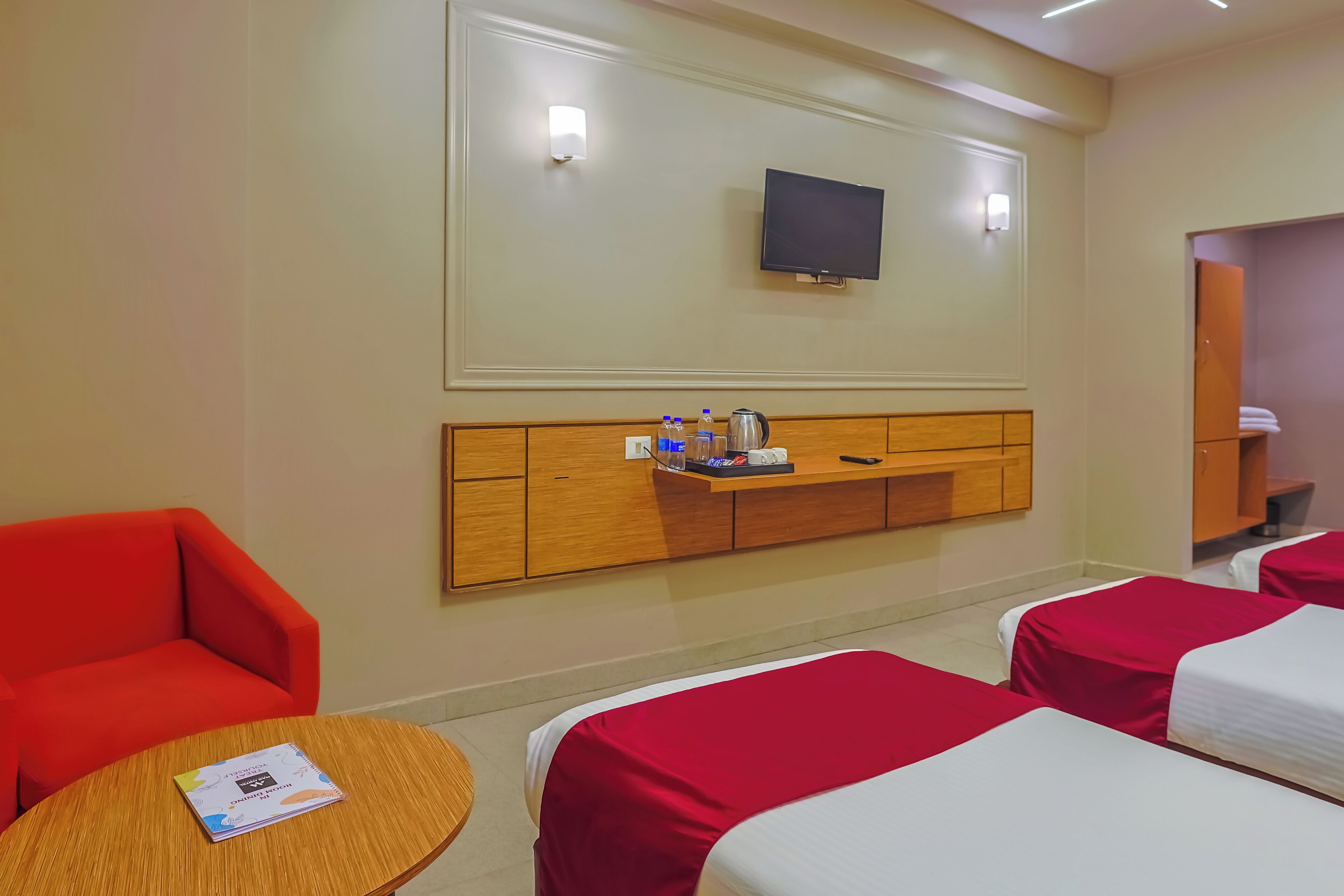 Habitación de hotel con dos camas, un televisor montado en la pared, cabecero de madera, una mesita con un libro y una silla roja.