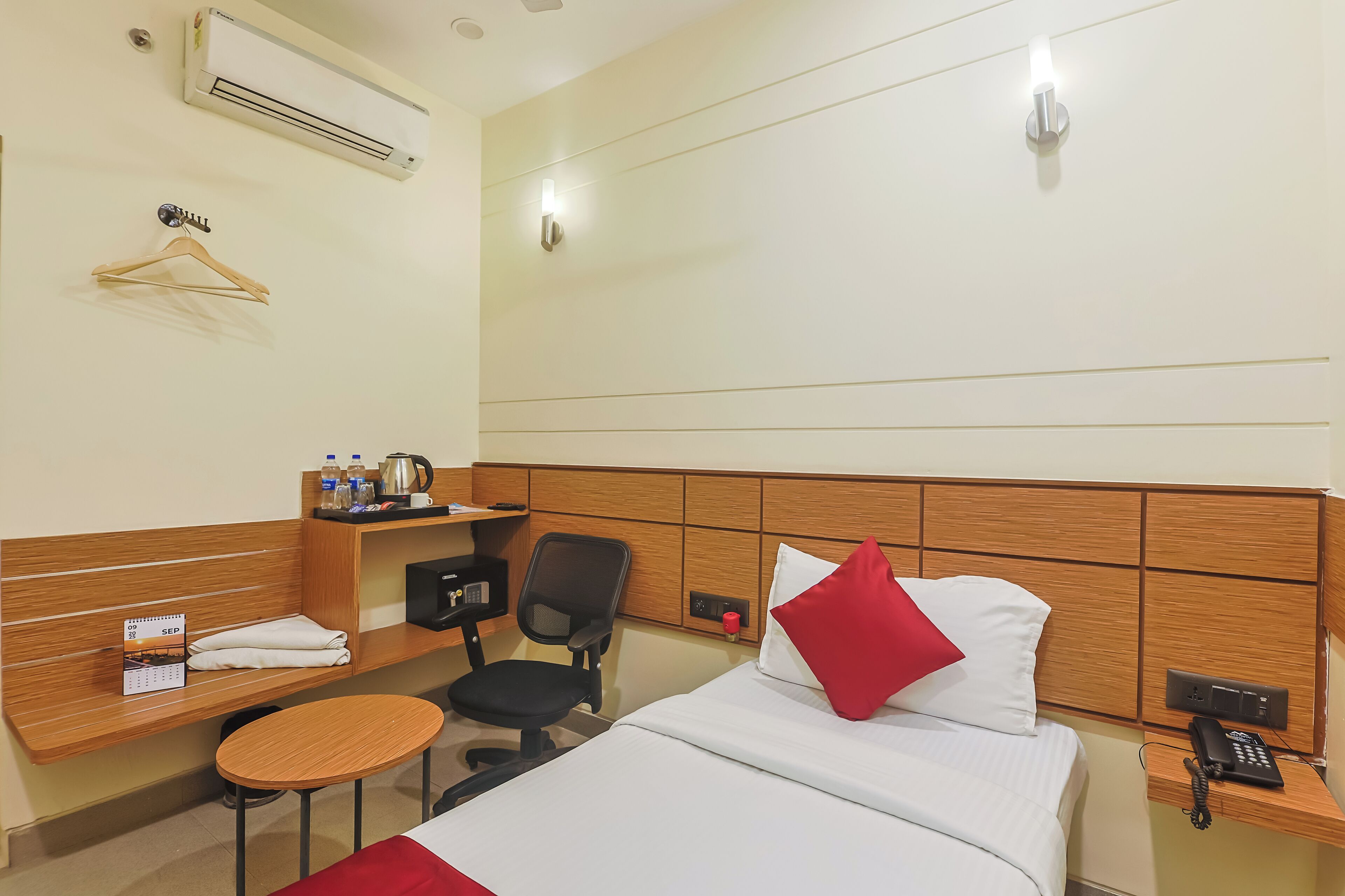 A hotel room with a bed, a desk with a chair, a small table, a shelf with toiletries, a calendar, and a phone on the bedside table.