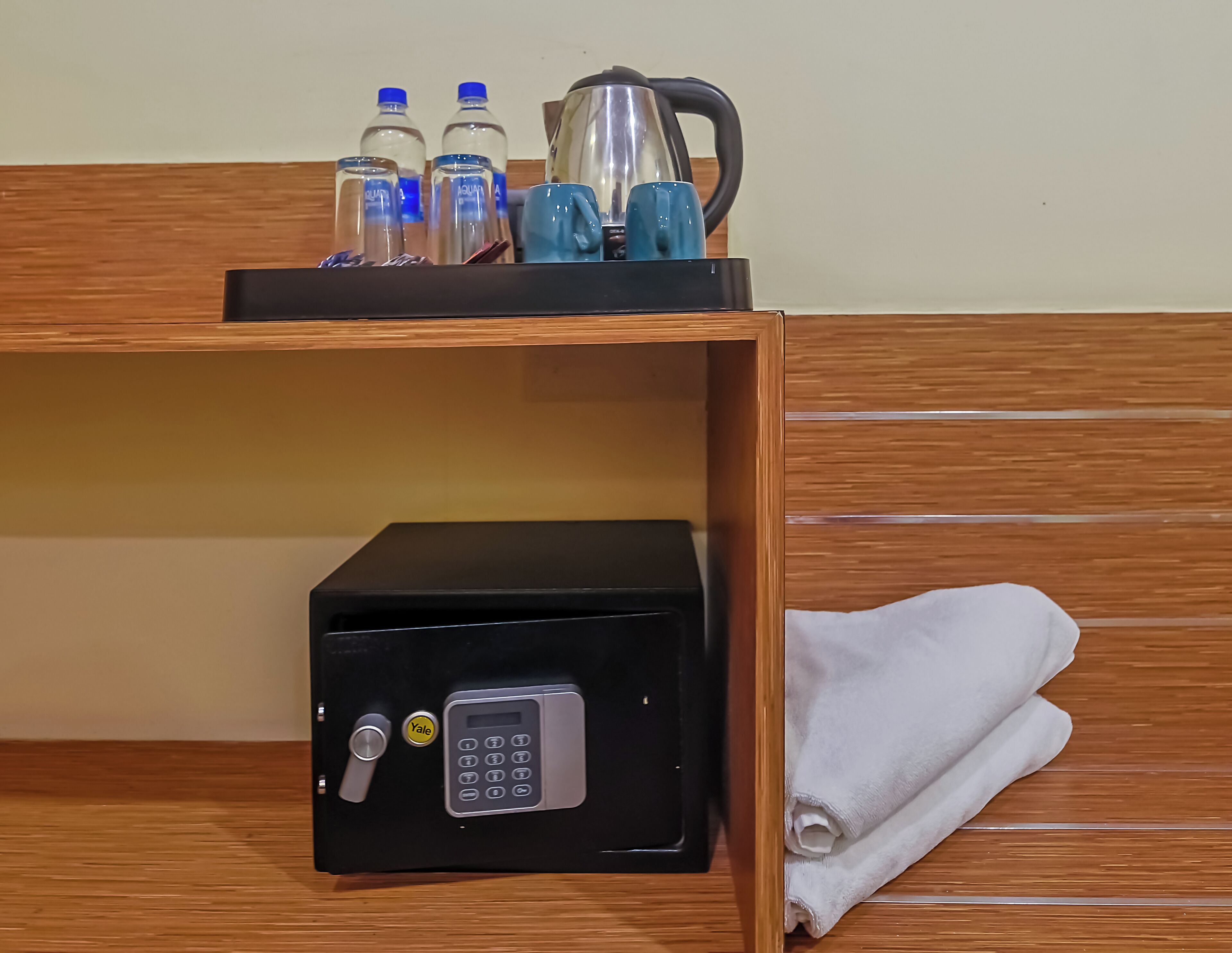 Habitación de hotel con una alacena minibar que contiene agua embotellada, una tetera y una cafetera. Debajo de la alacena, hay una caja fuerte con cerradura Yale.
