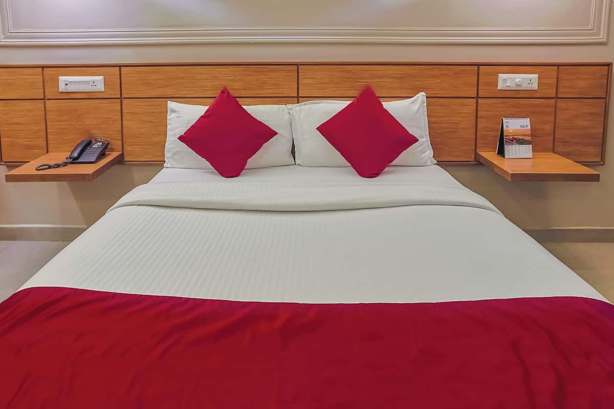 Una habitación de hotel con una cama, dos almohadas rojas, una colcha blanca, un edredón rojo, una mesita de noche con un teléfono y un calendario.