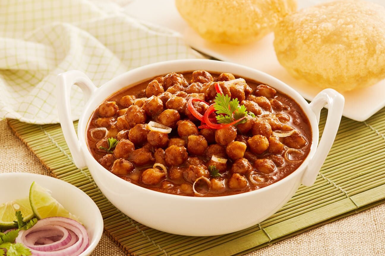 A bowl of chickpea curry with sliced onions and a red chili, accompanied by a plate of bread and a small bowl of sliced onions and cilantro.