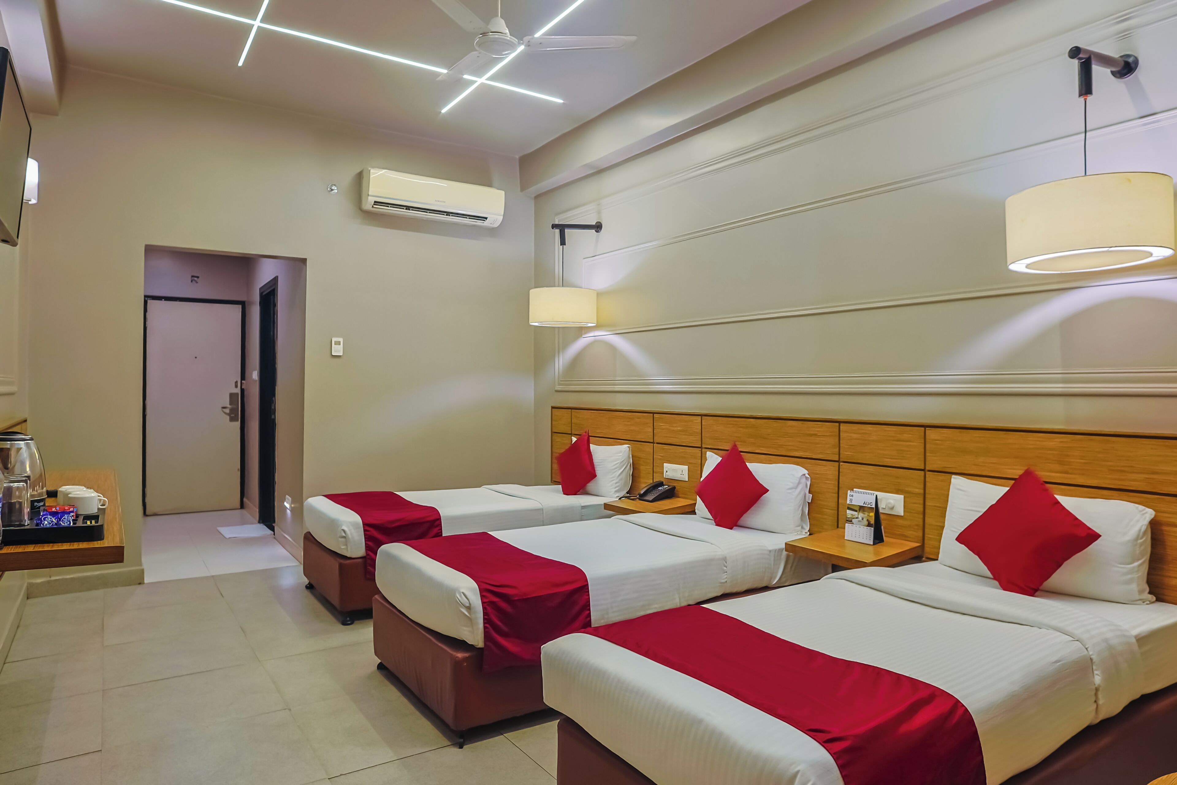 Habitación de hotel con tres camas, cada una con ropa de cama blanca y detalles rojos, cabecero de madera, mesita de noche con lámpara y ventilador de techo.