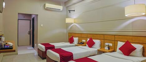 Habitación de hotel con tres camas, cada una con ropa de cama blanca y detalles rojos, cabecero de madera, mesita de noche con lámpara y ventilador de techo.
