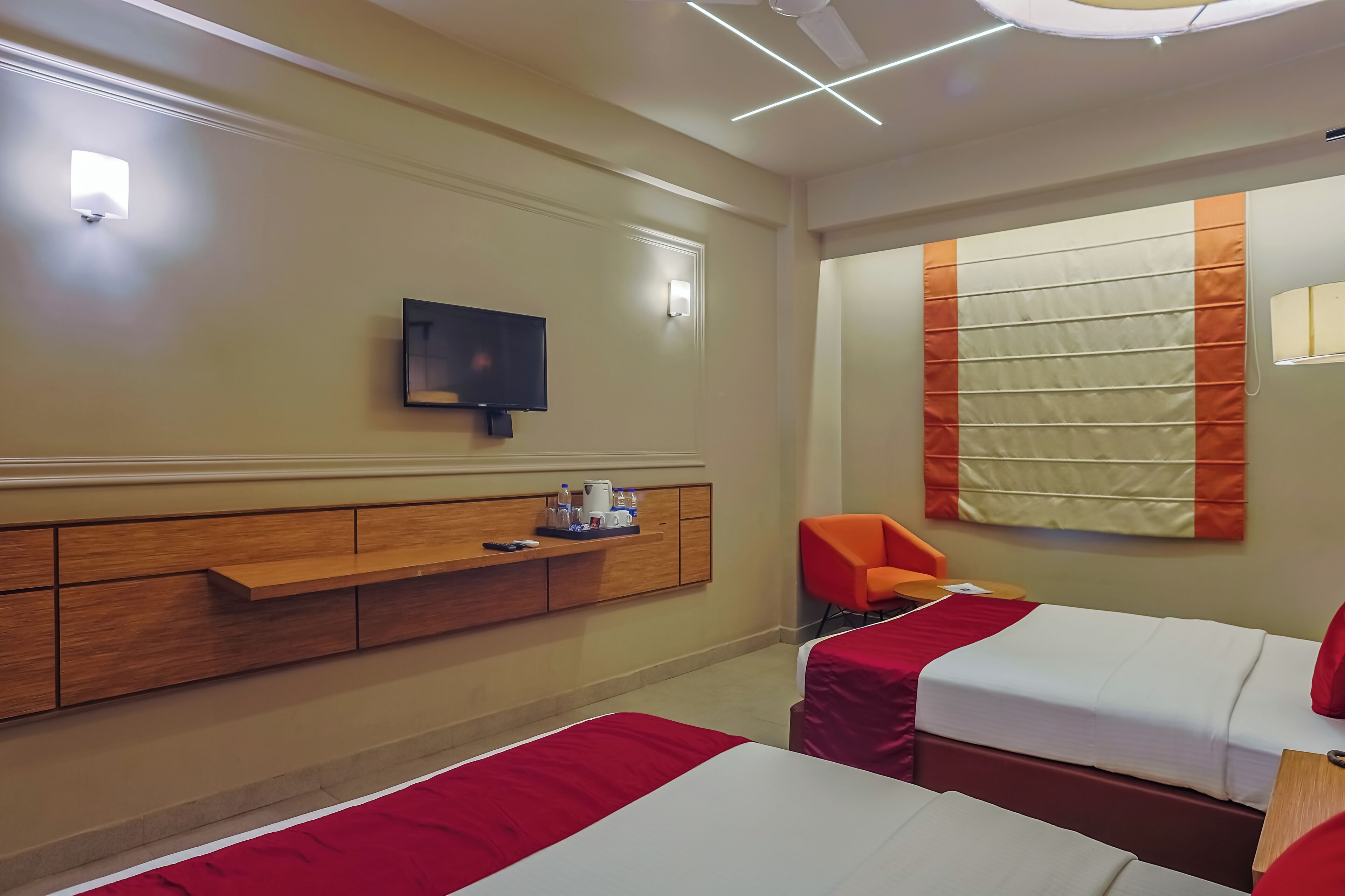 A hotel room with two beds, a TV, a desk with a coffee maker, and a large window with blinds.