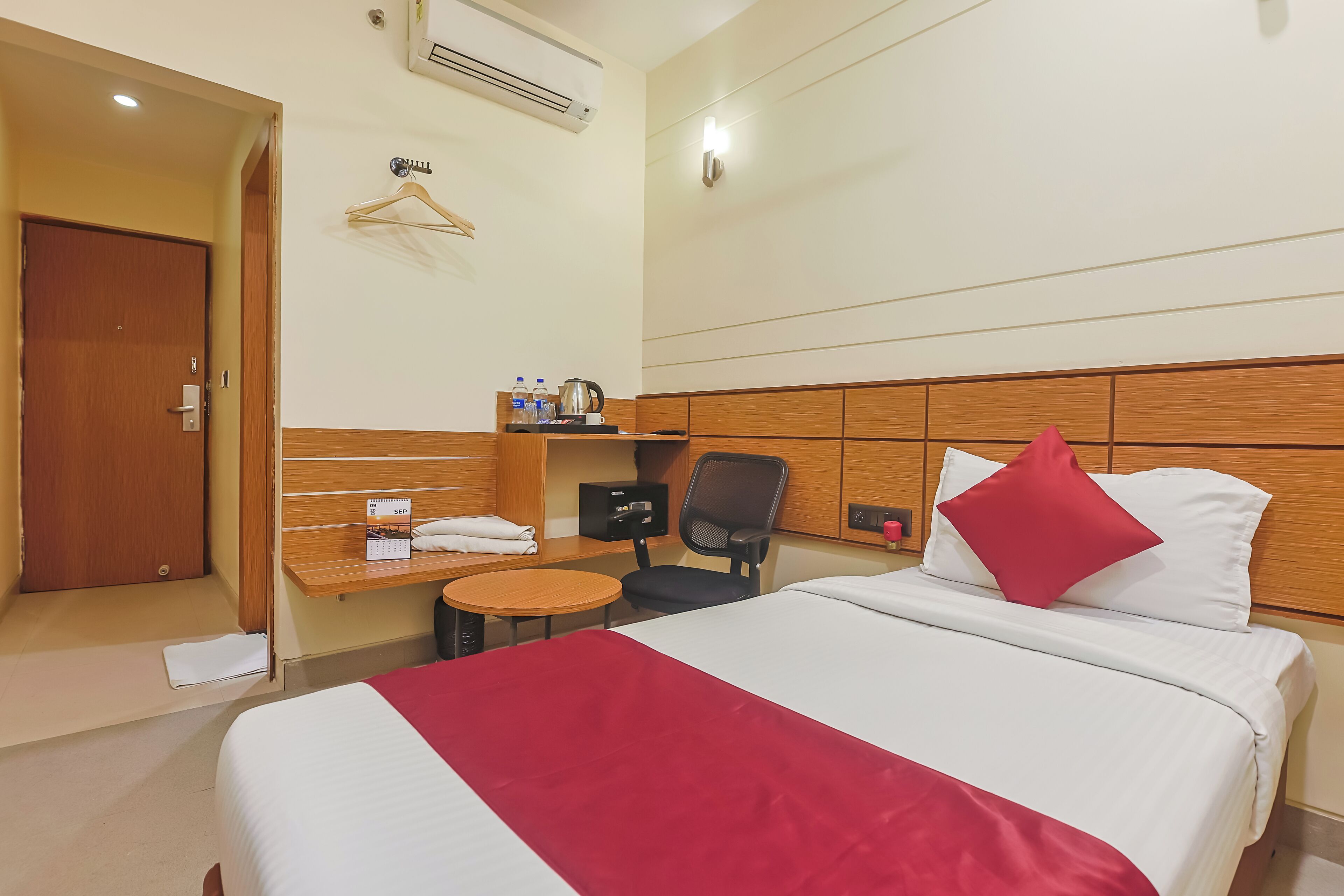 A hotel room with a bed, a desk with a chair, a small table, a hanging coat rack, and a wall-mounted air conditioner.