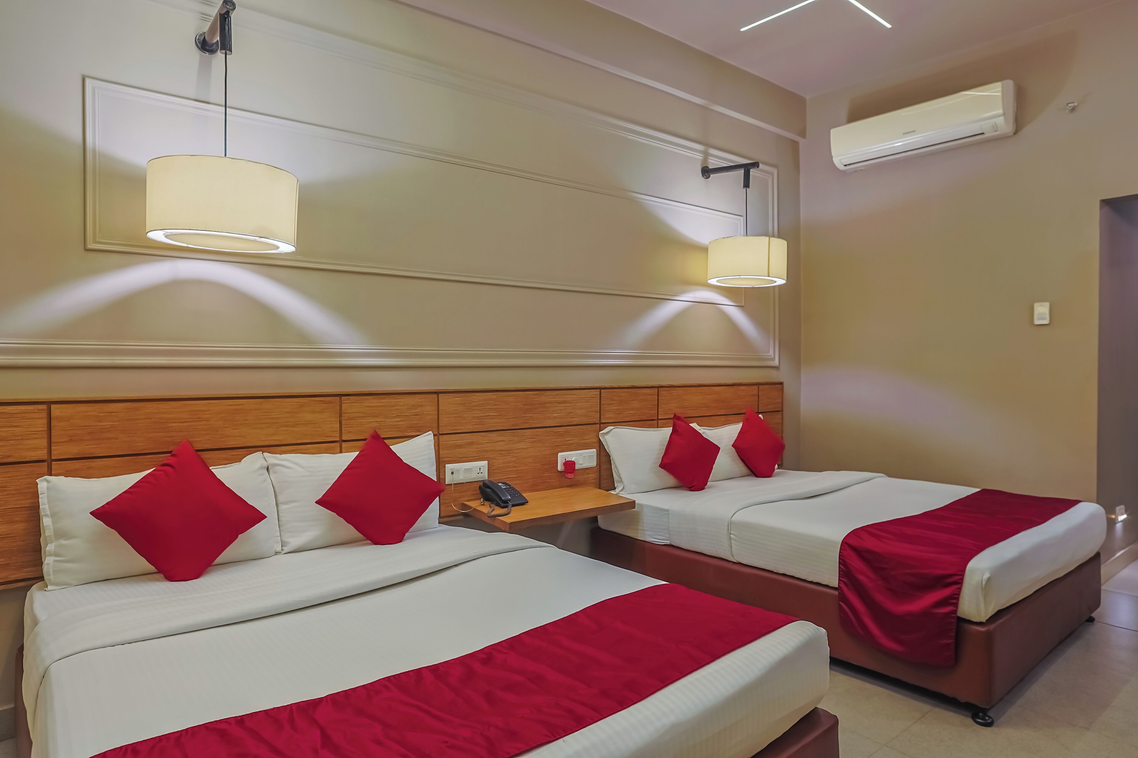 Hotelkamer met twee bedden, elk voorzien van witte lakens en rode kussens, een houten hoofdbord, een nachtkastje met een telefoon en een wandairconditioner.