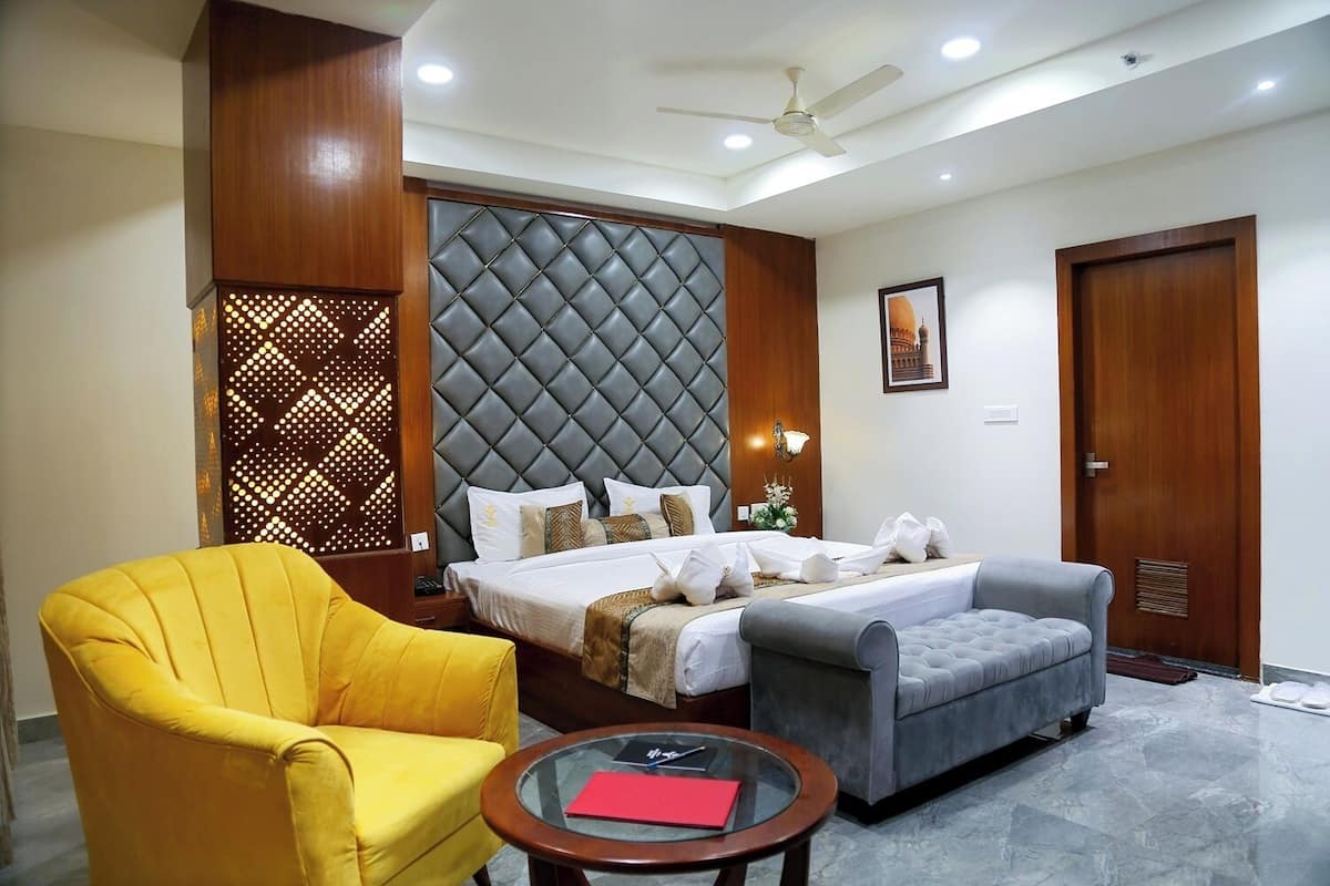 A hotel room with a large bed, a yellow armchair, a grey sofa, a glass coffee table, and a ceiling fan.