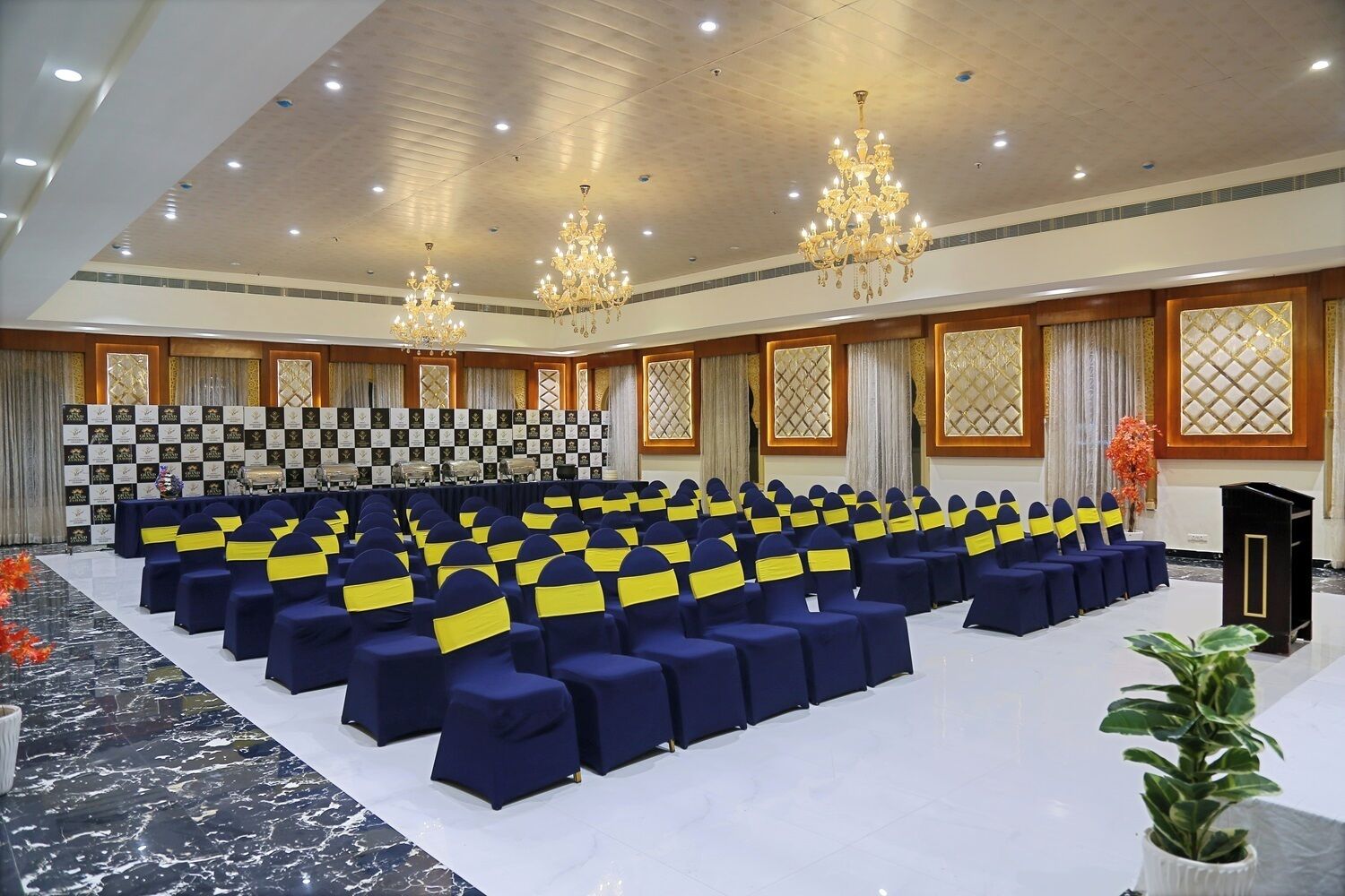 A conference room with rows of chairs, a stage with a checkered backdrop, chandeliers, and a potted plant.