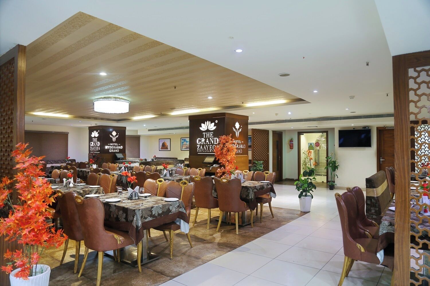 A spacious restaurant with multiple tables and chairs, a wooden ceiling, and decorative plants.