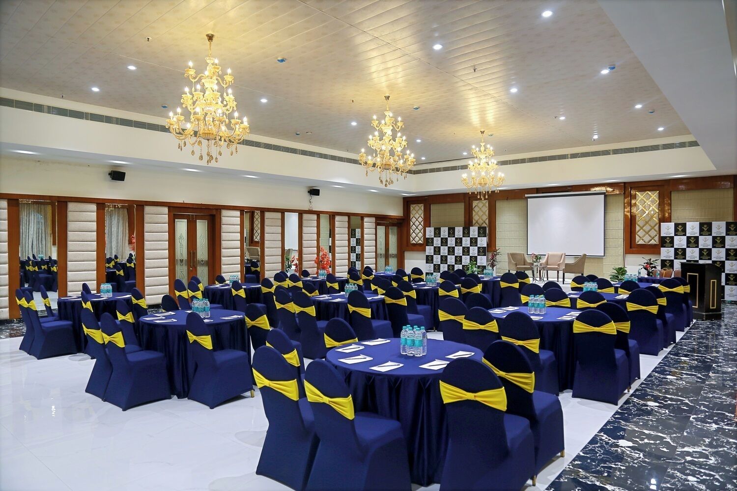 Een vergaderzaal met ronde tafels, marineblauwe stoelen met gele linten, kroonluchters en een projectiescherm.
