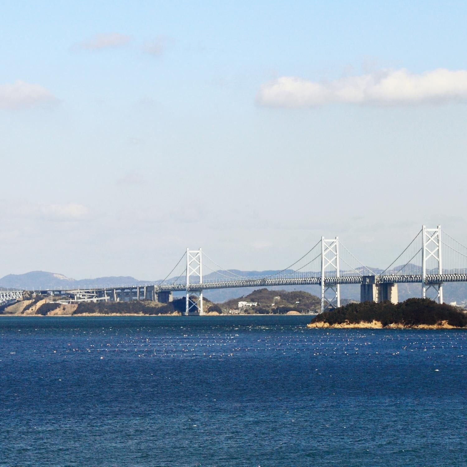 Eine große Hängebrücke überspannt ein Gewässer, im Hintergrund sind Hügel zu sehen.