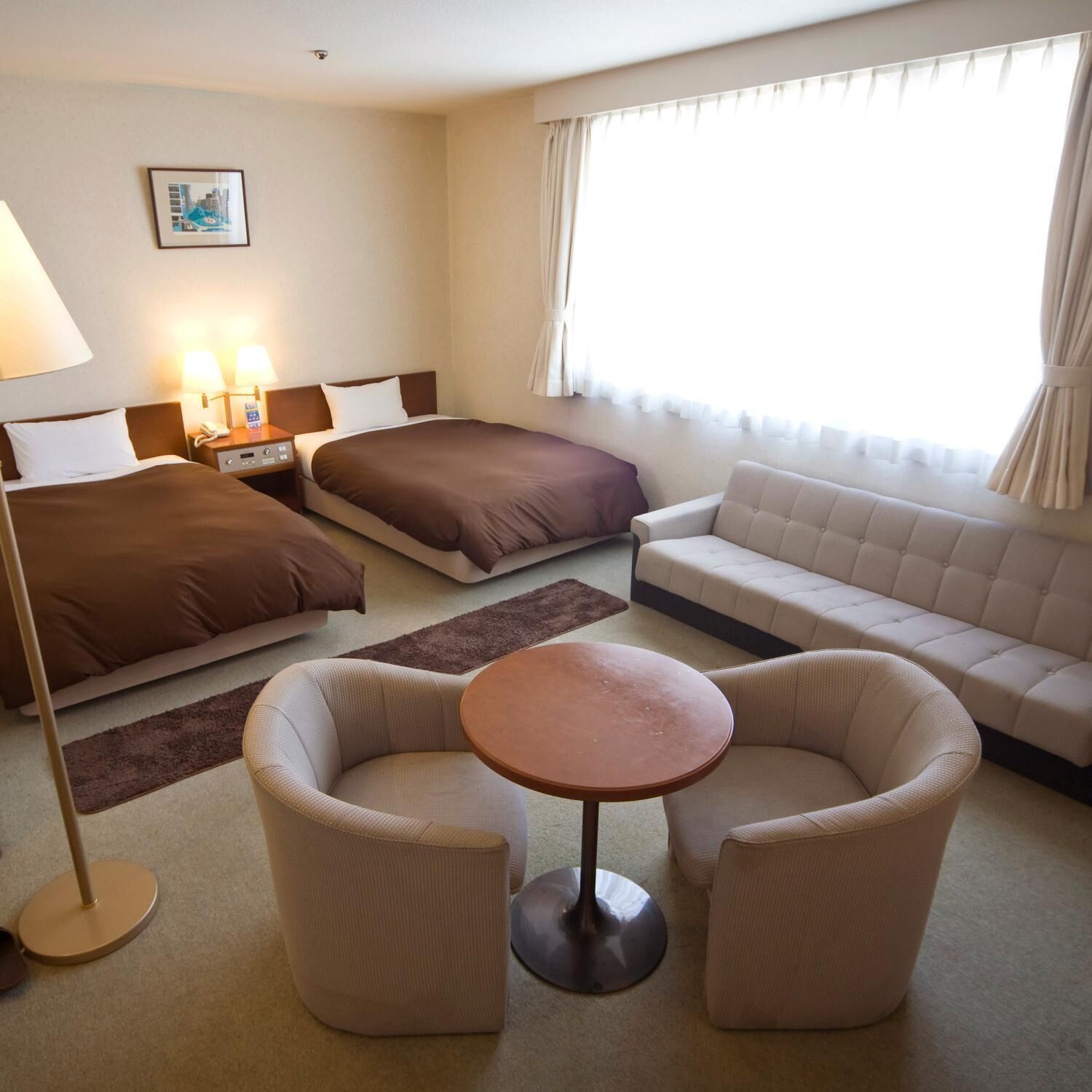 Ein Hotelzimmer mit zwei Betten, einem Sofa, zwei Sesseln, einem kleinen Tisch, einer Lampe und einem Bild an der Wand.
