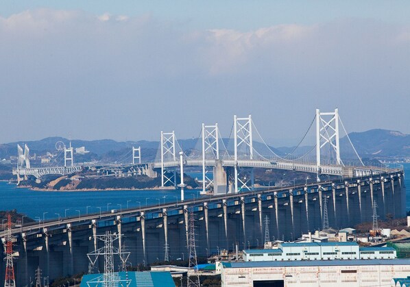 A large suspension bridge with multiple towers and cables, spanning a body of water with a hilly landscape in the background.