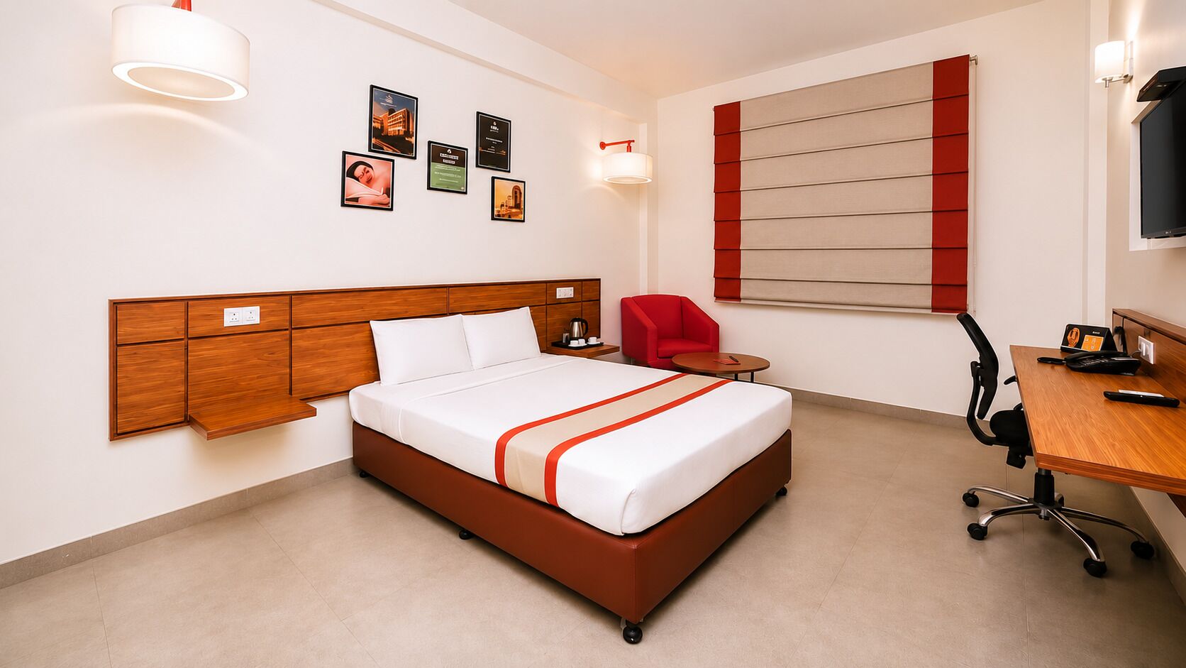 A hotel room with a wooden headboard, a bed with white bedding and red accents, a desk with a chair, a red armchair, and wall-mounted artwork.