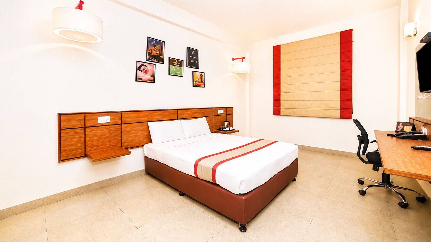A hotel room with a bed, bedside table, lamp, wall-mounted artwork, and a desk with a chair.