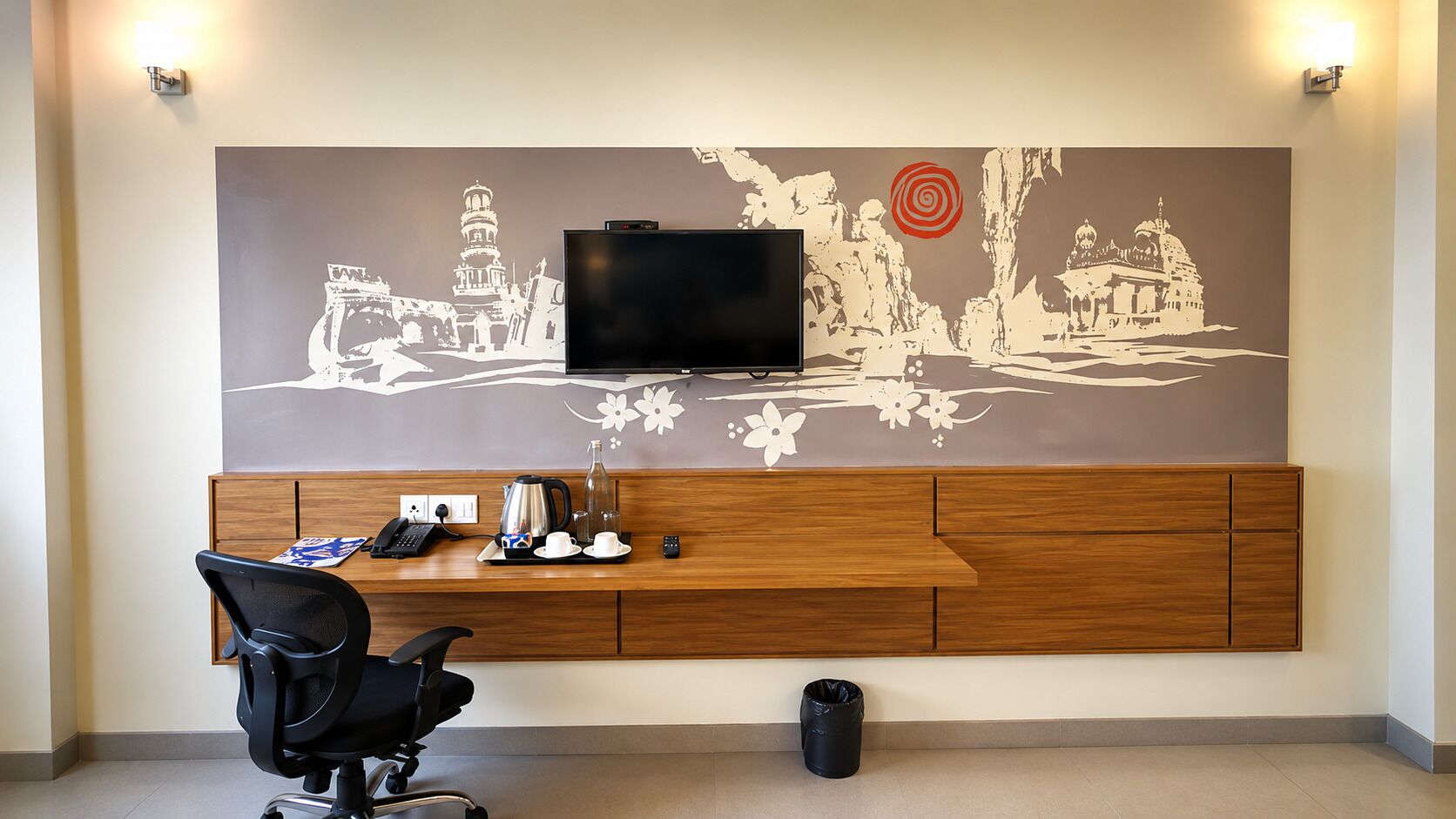 A hotel room with a flat-screen TV mounted on a wall with a decorative mural, a wooden desk with a chair, and a trash bin.