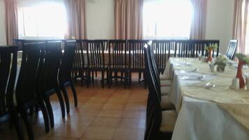 Un comedor con sillas y mesas negras, ventanas amplias con cortinas y piso de baldosas.
