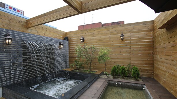 Una terraza de madera con un jacuzzi y un pequeño estanque, rodeada de paredes de madera y un muro de ladrillo con una cascada.