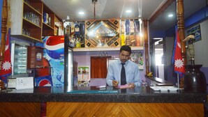 A reception desk with a man writing, surrounded by shelves with various items, a calendar, and a decorative wall. - Hotel Deep Sagar (Pokhara)