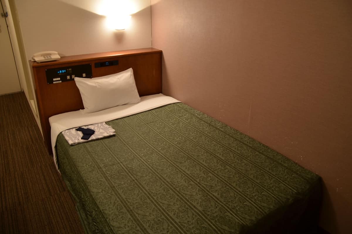 Quarto de hotel com cama, telefone de apoio e luminária fixada na parede.