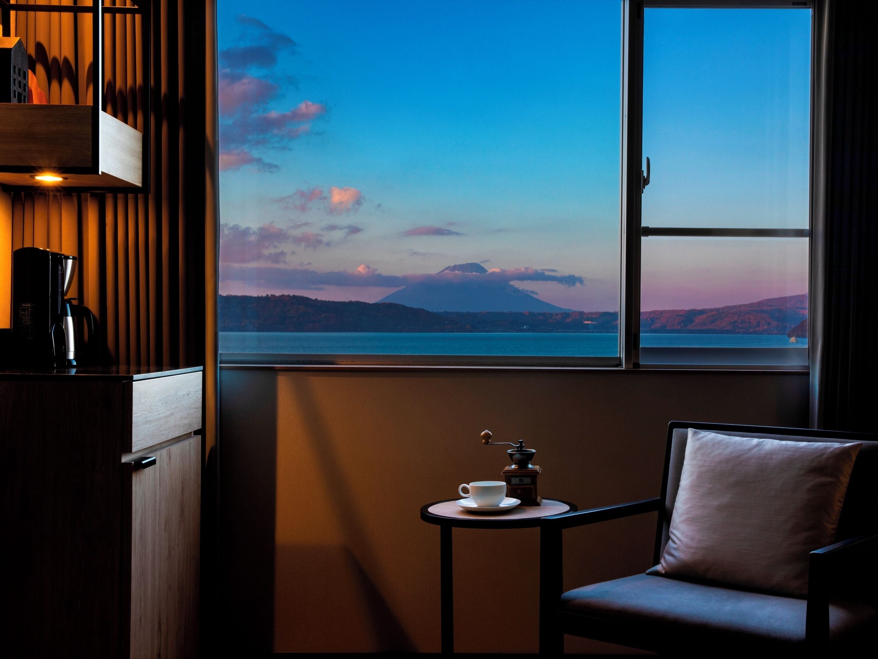 A cozy room with a view of a lake and mountains, a small table with a coffee pot and cup, and a chair.