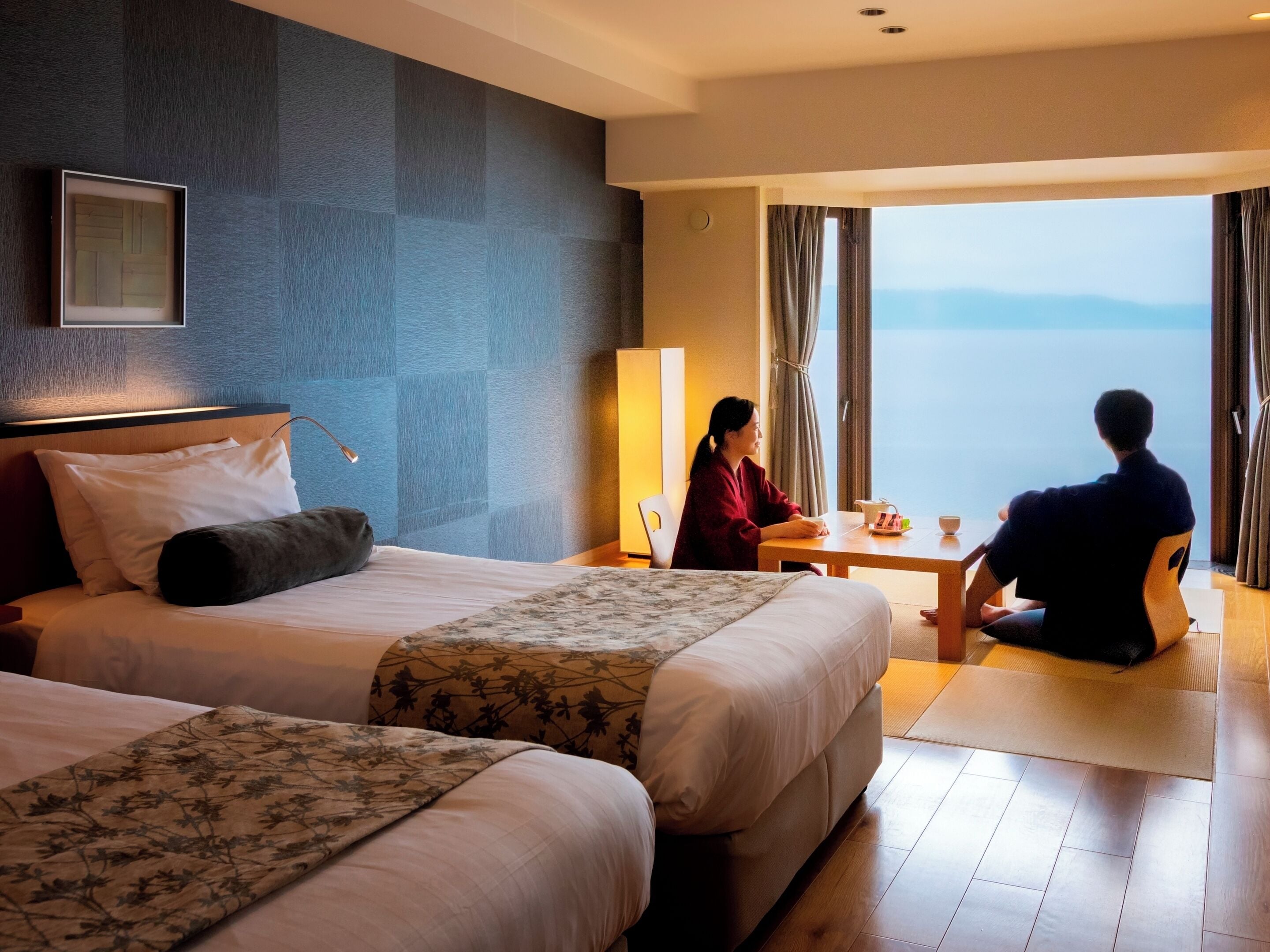 A hotel room with two beds, a sitting area with a table, and a view of the sea.