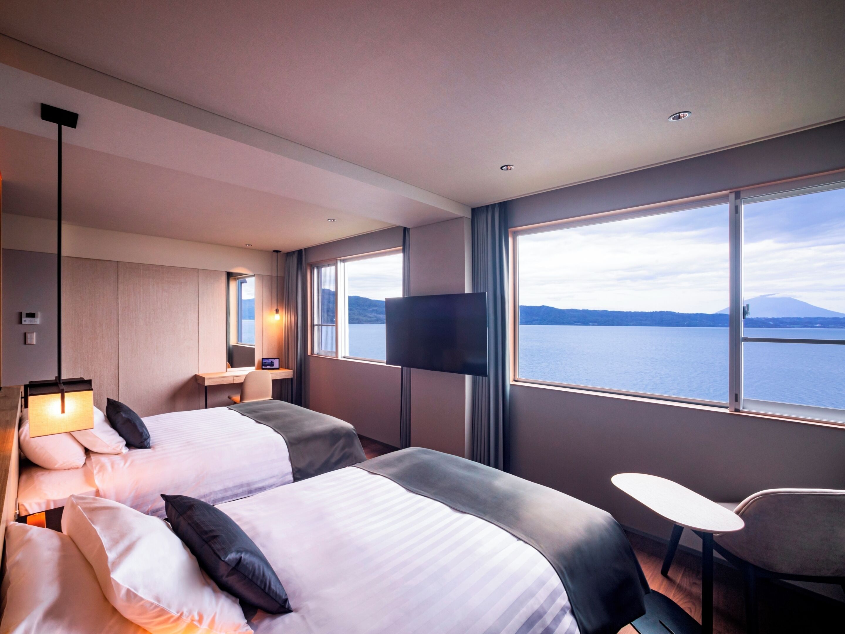 A hotel room with two beds, a desk, a TV, and a view of a body of water.