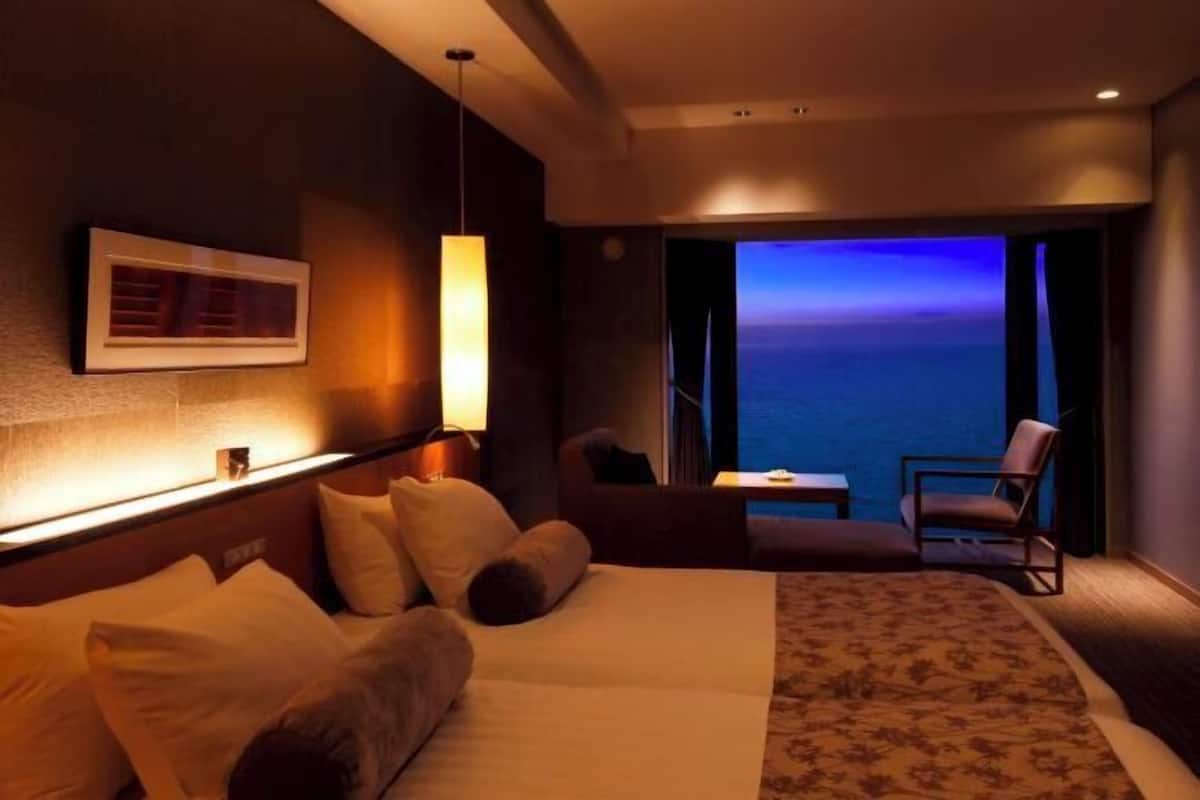 Une chambre d’hôtel avec un lit, une chaise, une petite table, une vue sur l’océan et un tableau encadré au mur.