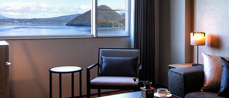 A hotel room with a view of a lake and mountains, a chair, a small table with a cup and a book, and a lamp.