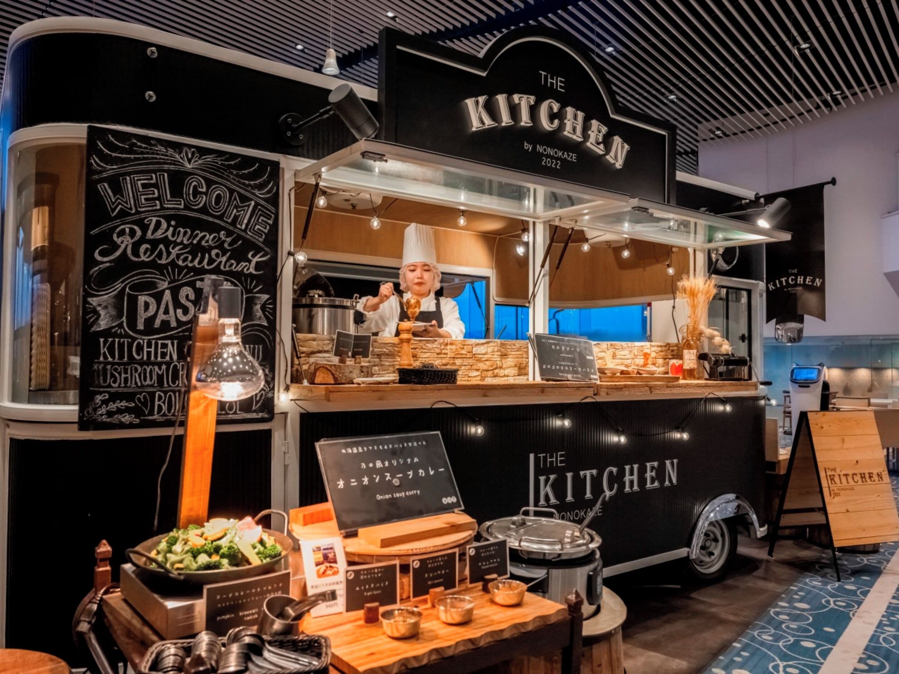 A food truck named "The Kitchen" with a chef preparing food, a signboard with menu items, and various cooking utensils displayed.