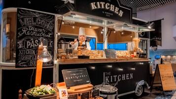 A food truck named "The Kitchen" with a chef preparing food, a signboard with menu items, and various cooking utensils displayed.