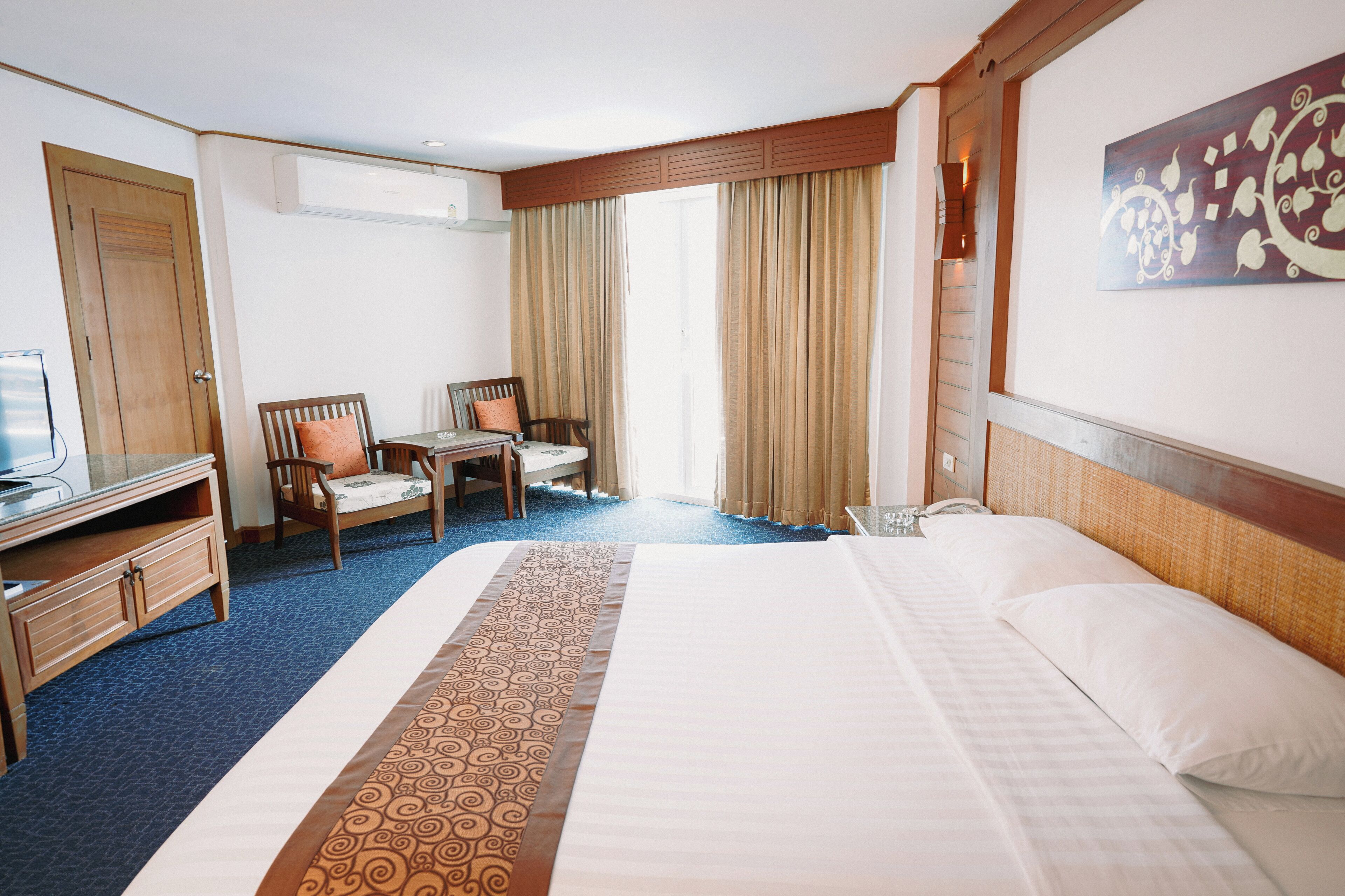 A hotel room with a large bed, a desk, two chairs, a television, and a window with curtains.
