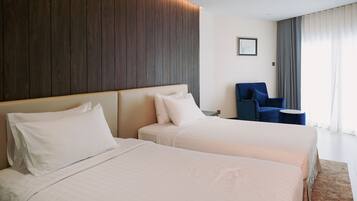 A hotel room with two beds, a blue armchair, and a large window with curtains.