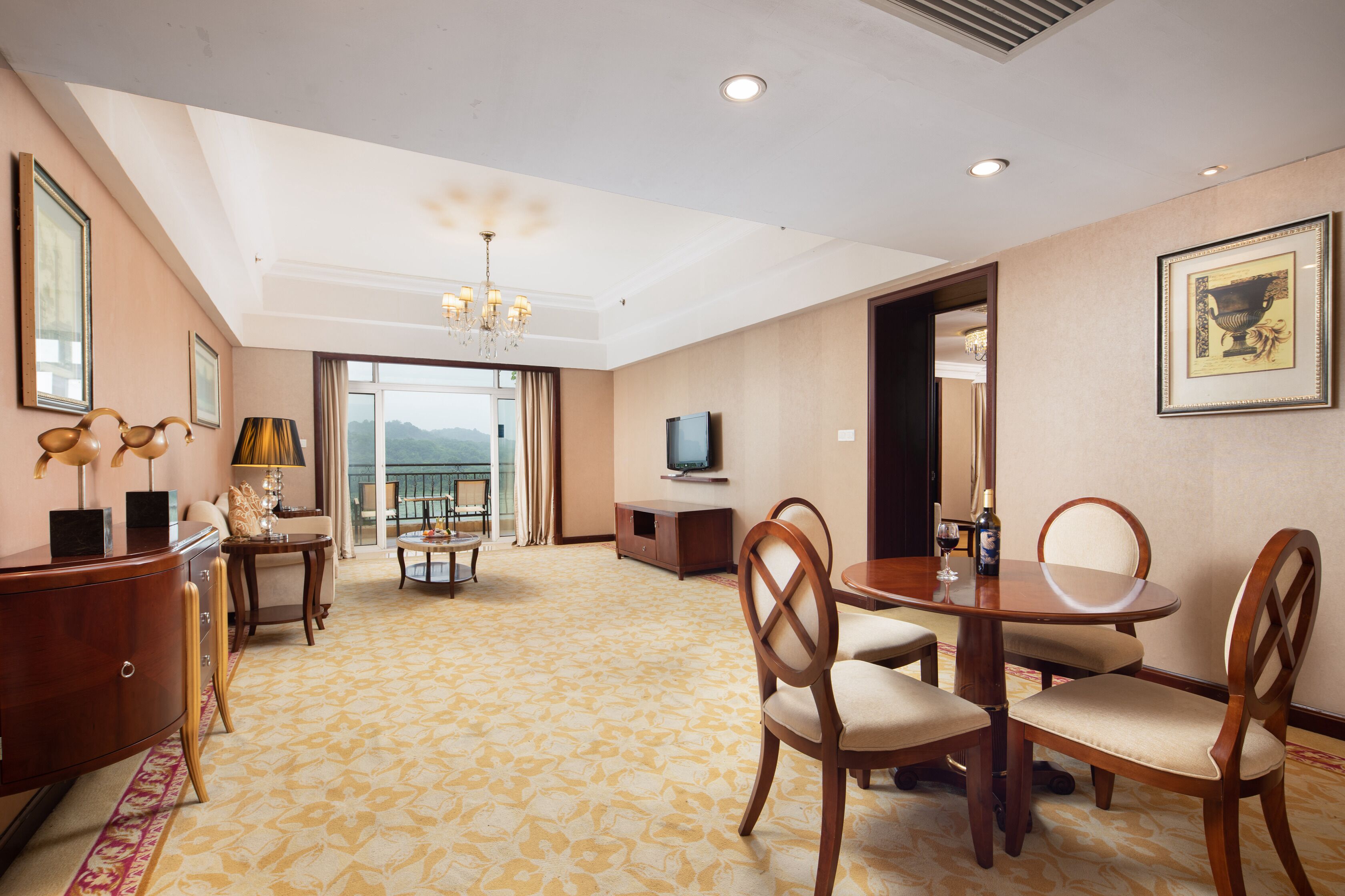 A hotel room with a dining area, a television, a chandelier, and a view of the outdoors.