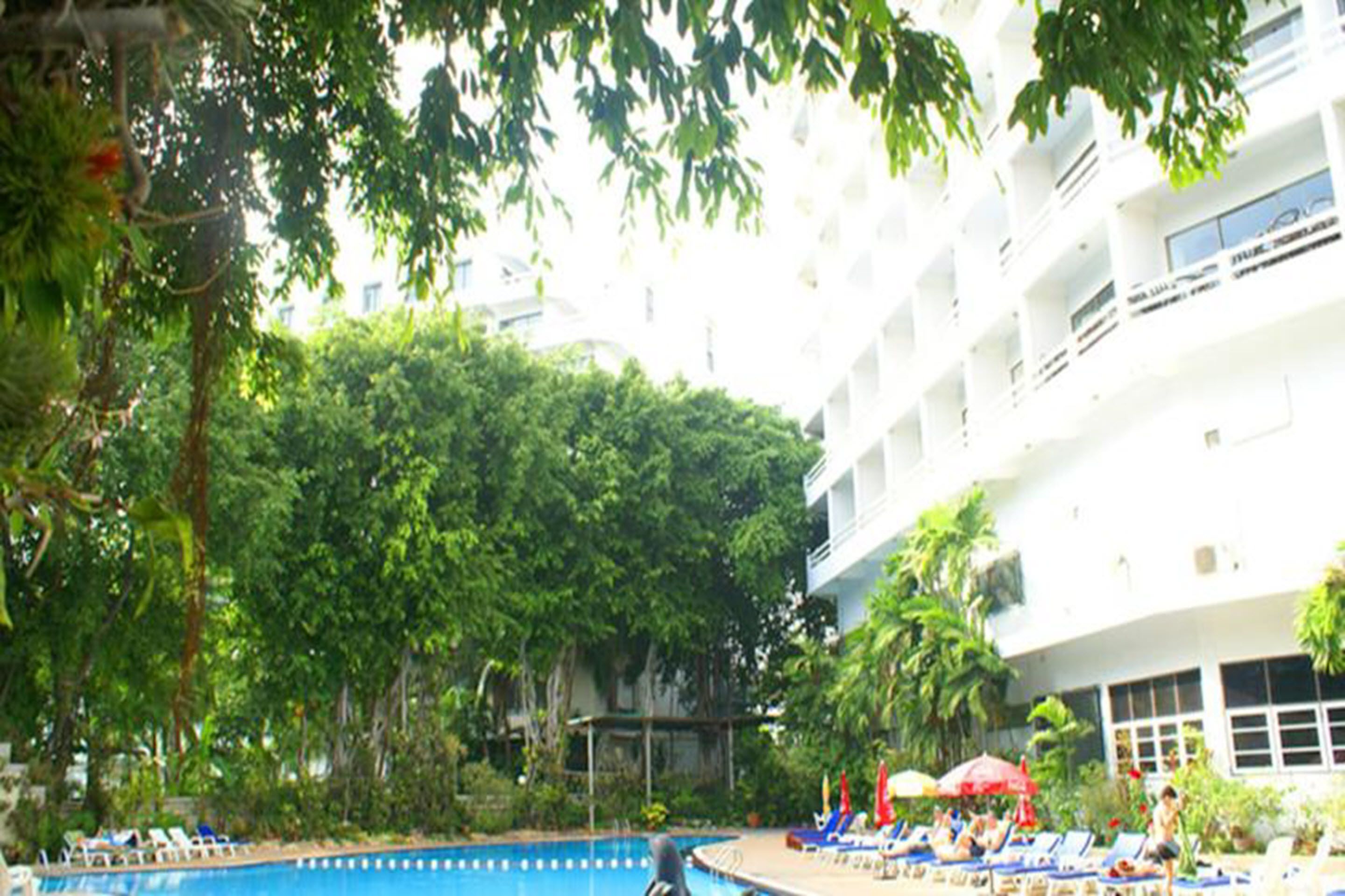 A pool area with lounge chairs and umbrellas, surrounded by lush greenery and a multi-story building.