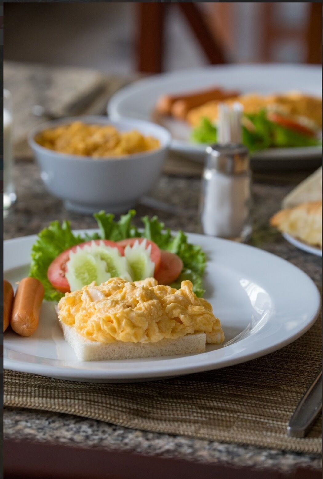 Une assiette avec des œufs brouillés sur des toasts, un hot-dog et une salade composée de laitue, de tomates et de concombres.
