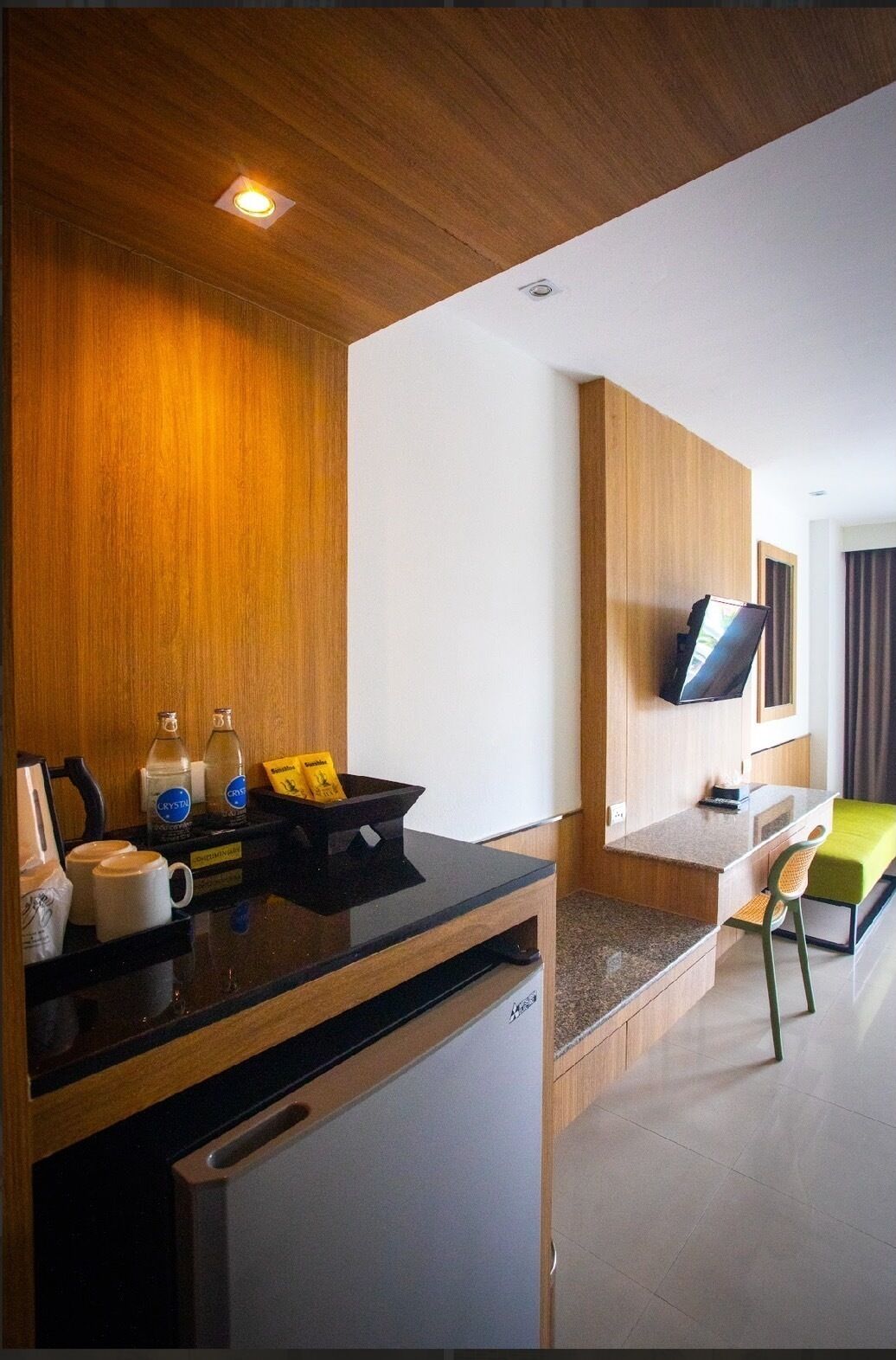 A modern hotel room with a wooden ceiling, a flat-screen TV mounted on the wall, a small kitchenette with a refrigerator, a dining area with a green chair, and a small table.