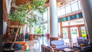 Un hall d’entrée spacieux avec de hauts plafonds, de grandes colonnes et un arbre au centre. On y trouve des espaces aménagés avec des tables et des chaises.