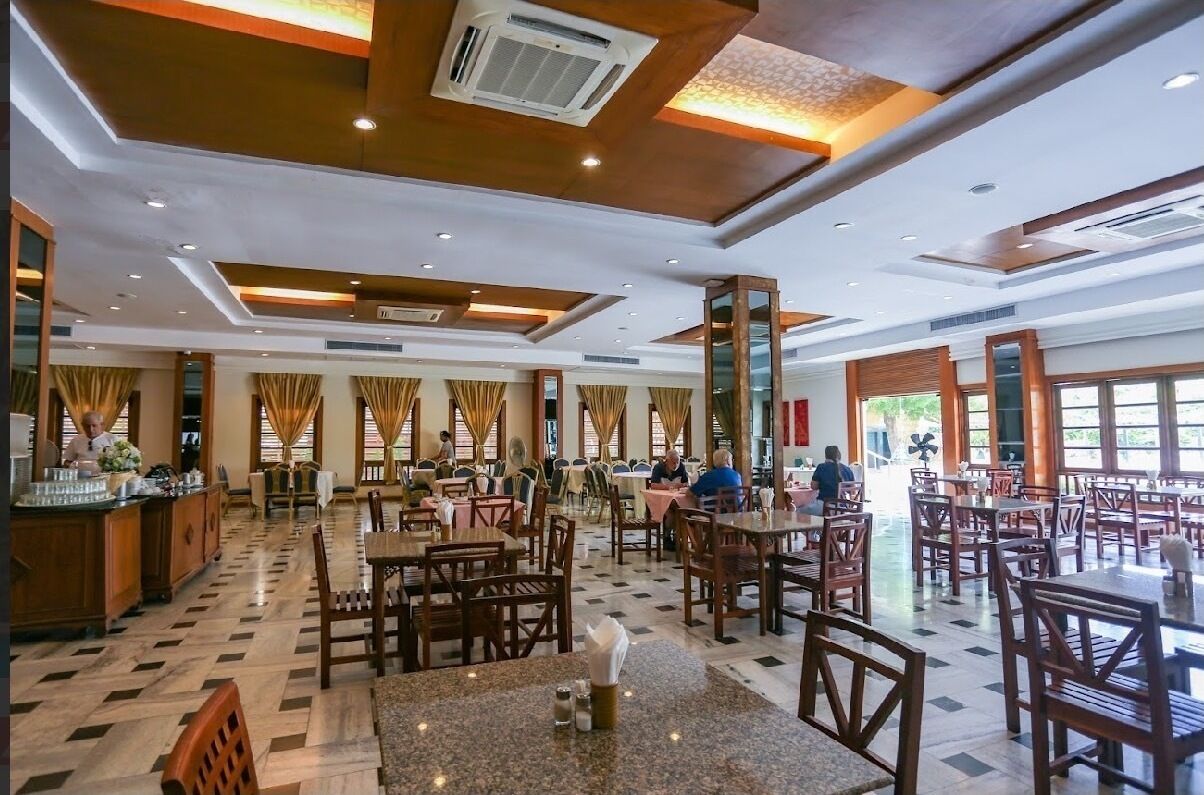 A spacious restaurant with multiple dining tables and chairs, a bar area, and large windows allowing natural light.