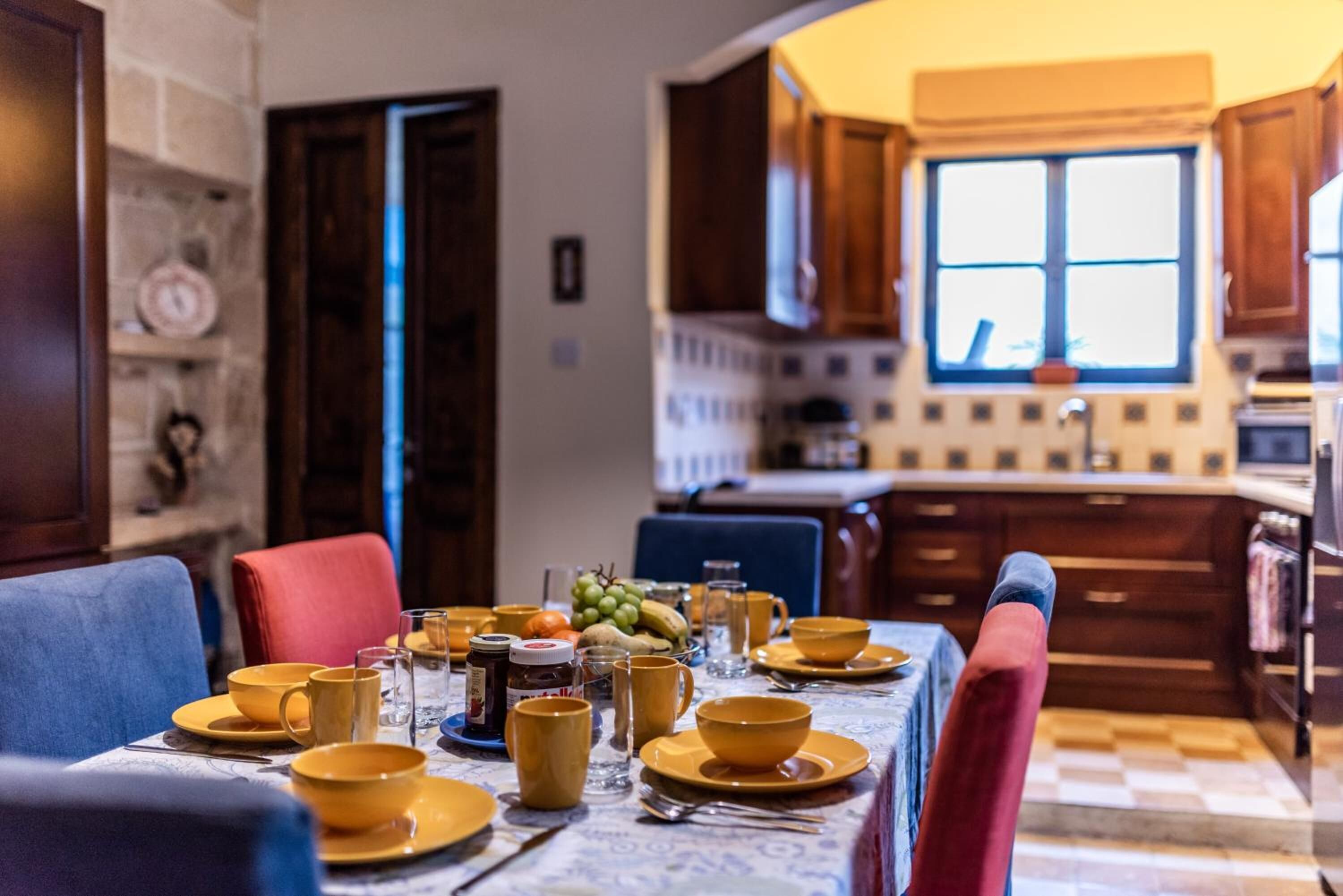 A dining area with a table set for four, surrounded by wooden chairs. A kitchen is visible in the background with a window above the sink.