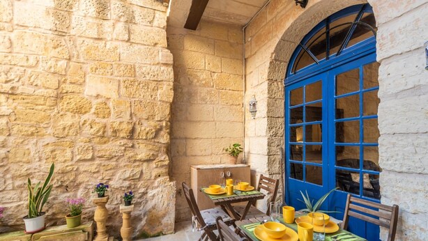 Zona pranzo all'aperto con pareti in pietra, una porta blu e arredi in legno.