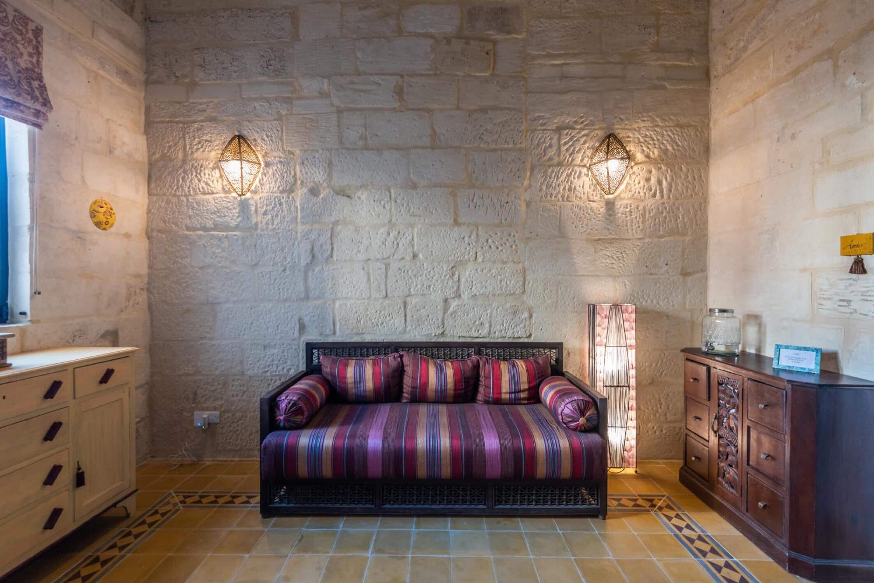 A room with a stone wall, a striped sofa, a wooden cabinet, and two wall-mounted lamps.