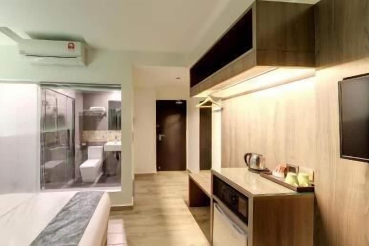 A modern hotel room with a bed, a TV, a kitchenette, and a bathroom with a glass shower.