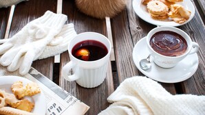 A wooden table with a plate of cookies, two cups of hot beverages, a knitted glove, and a fur pom-pom.