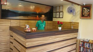 A reception desk with a wooden finish, a framed painting of a sunset, and a fan on the wall.