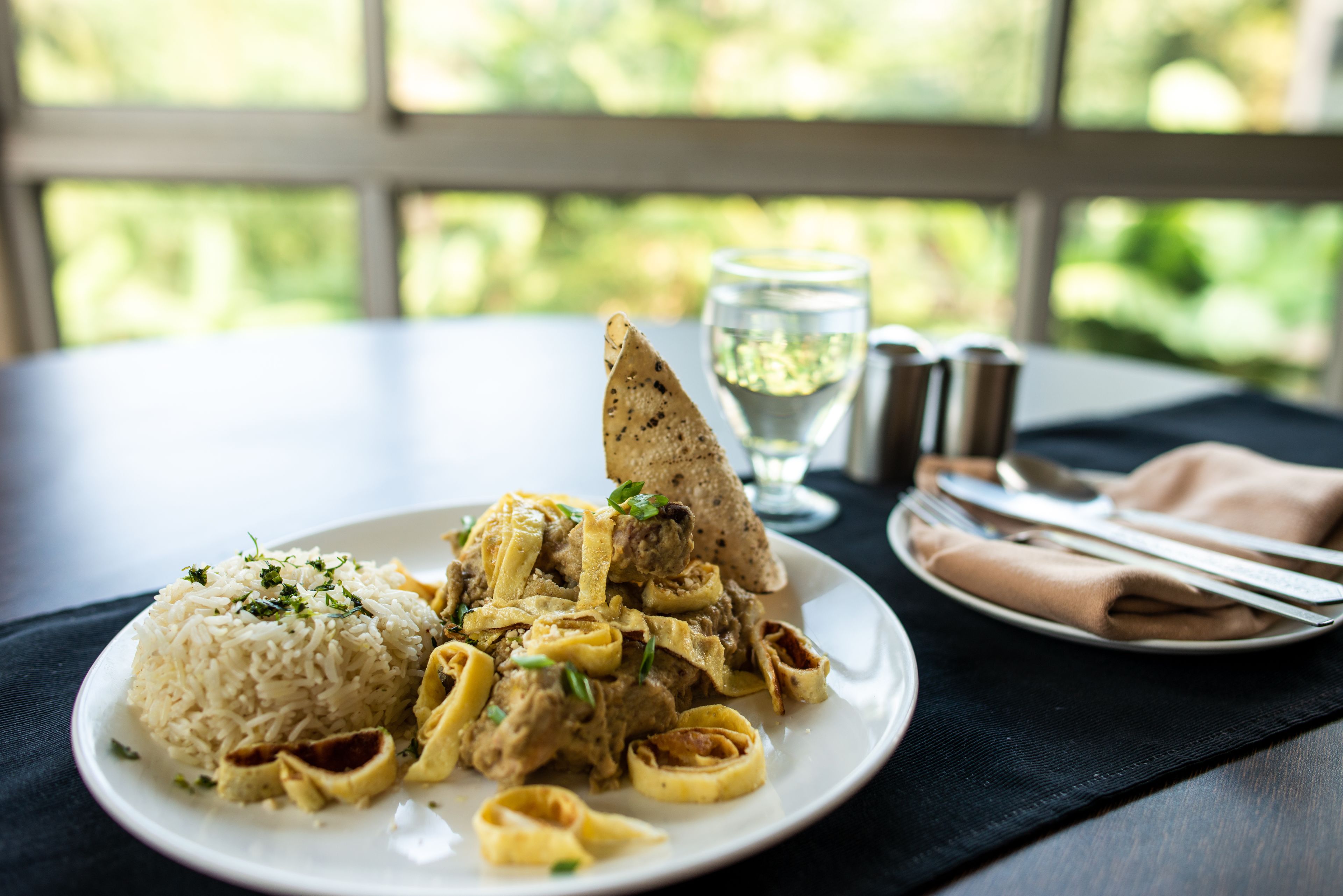 A plate of food with rice and pasta, a glass of water, and cutlery on a table with a view of greenery outside.