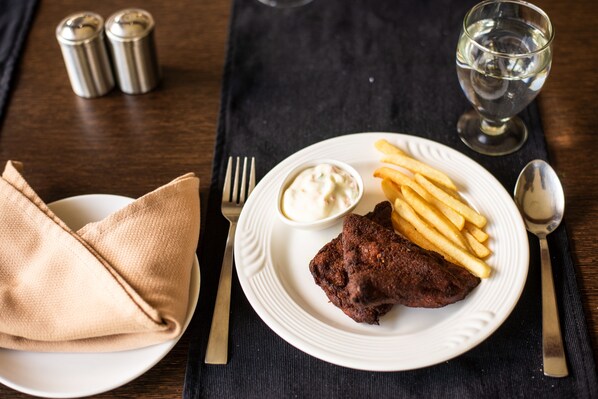 Um prato com bife, batatas fritas e uma pequena tigela de molho, acompanhado de um copo de água e saleiro e pimenteiro sobre uma mesa.