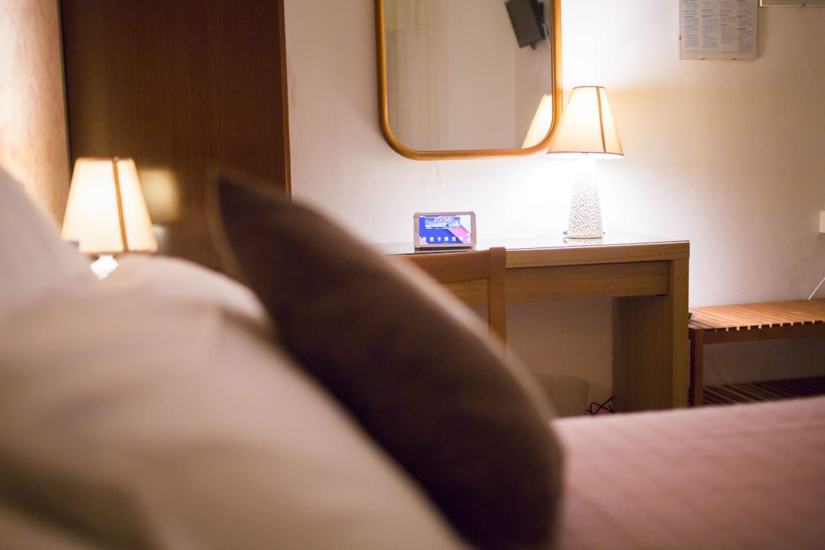 A hotel room with a bed, a bedside table with a lamp, and a small clock displaying the time.