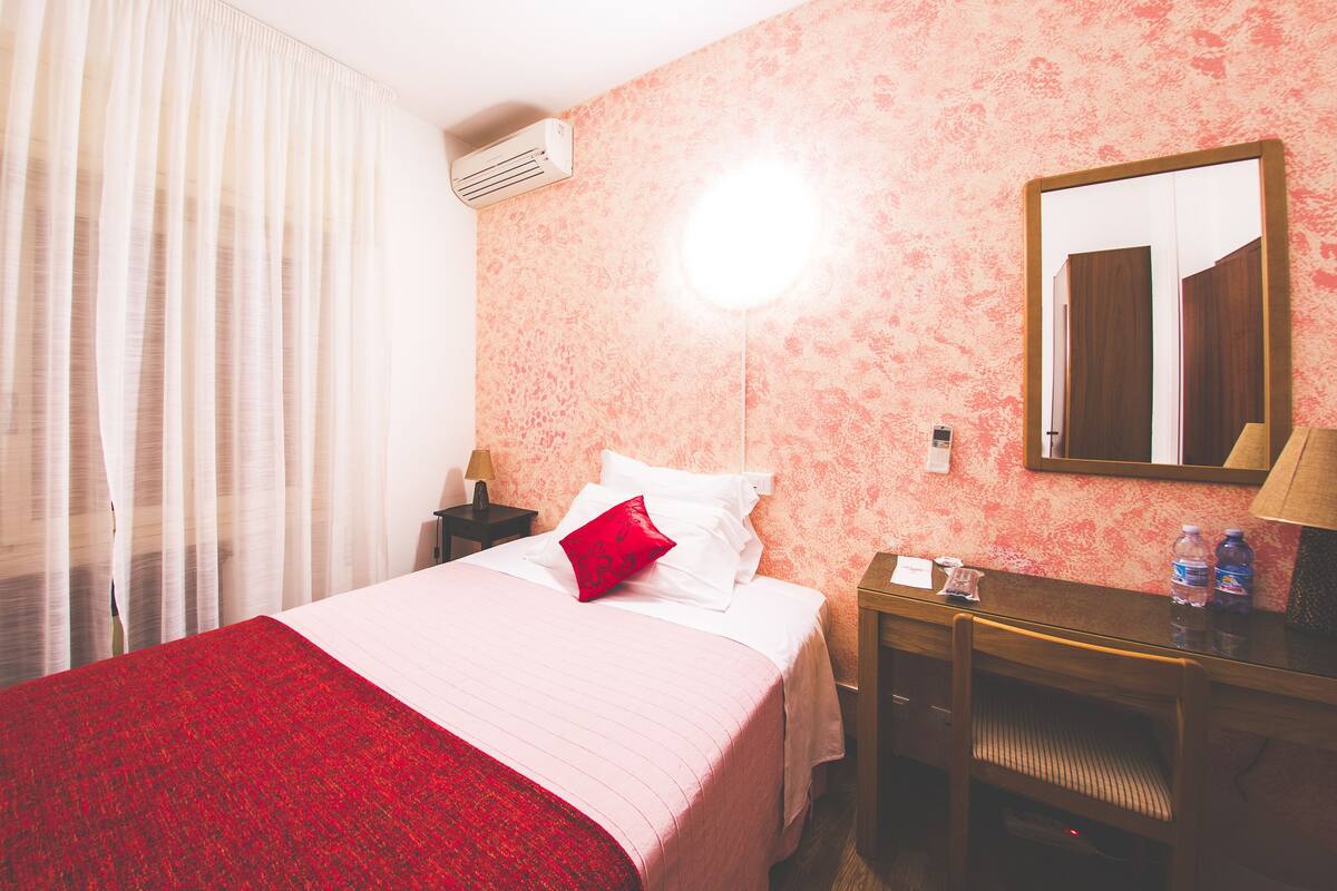 A hotel room with a bed, a desk, a mirror, and an air conditioning unit.
